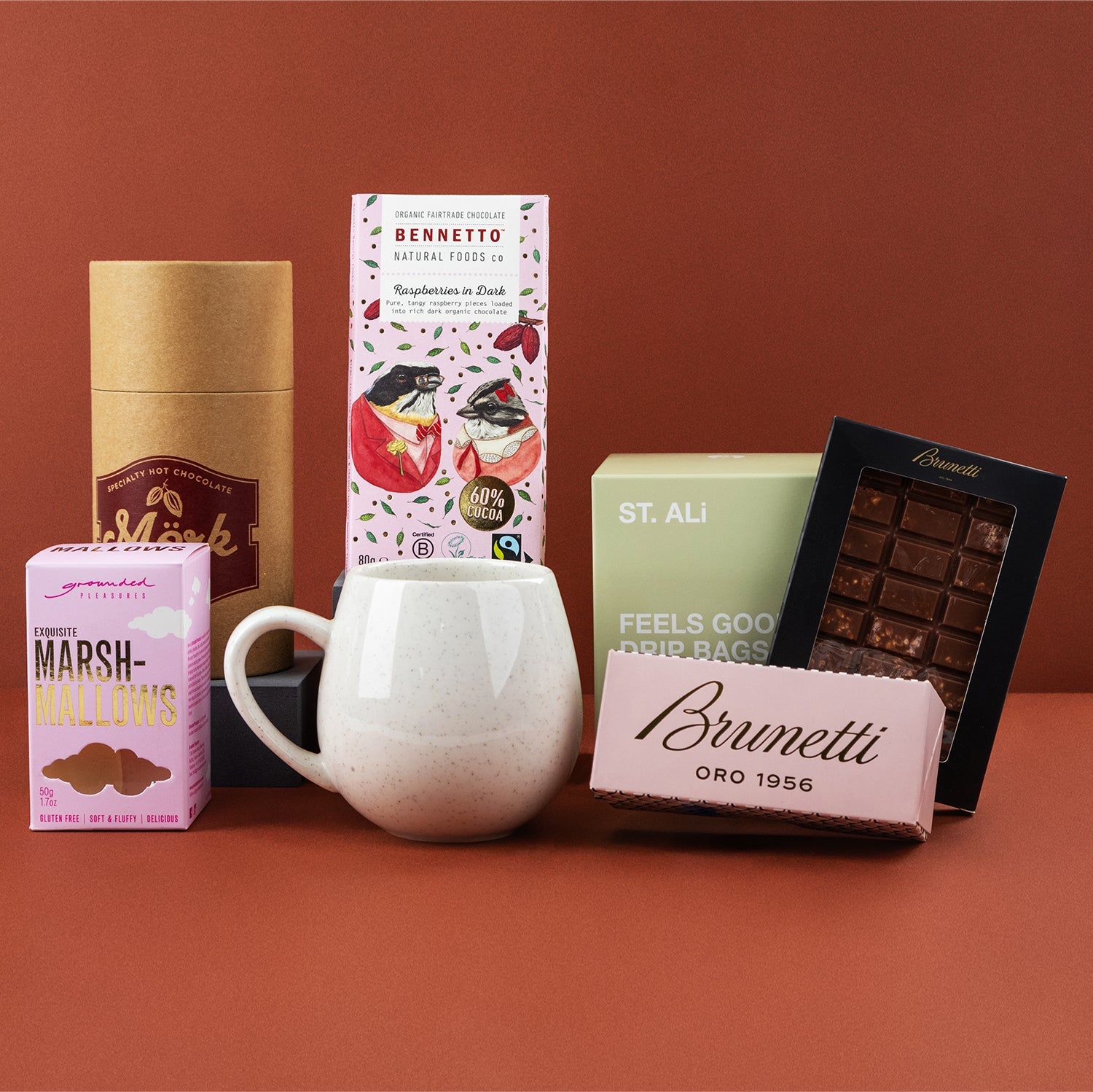 Coffee and Treats Hamper with white ceramic mug, pink Brunetti chocolate box, marshmallows, and gourmet snacks on brown.