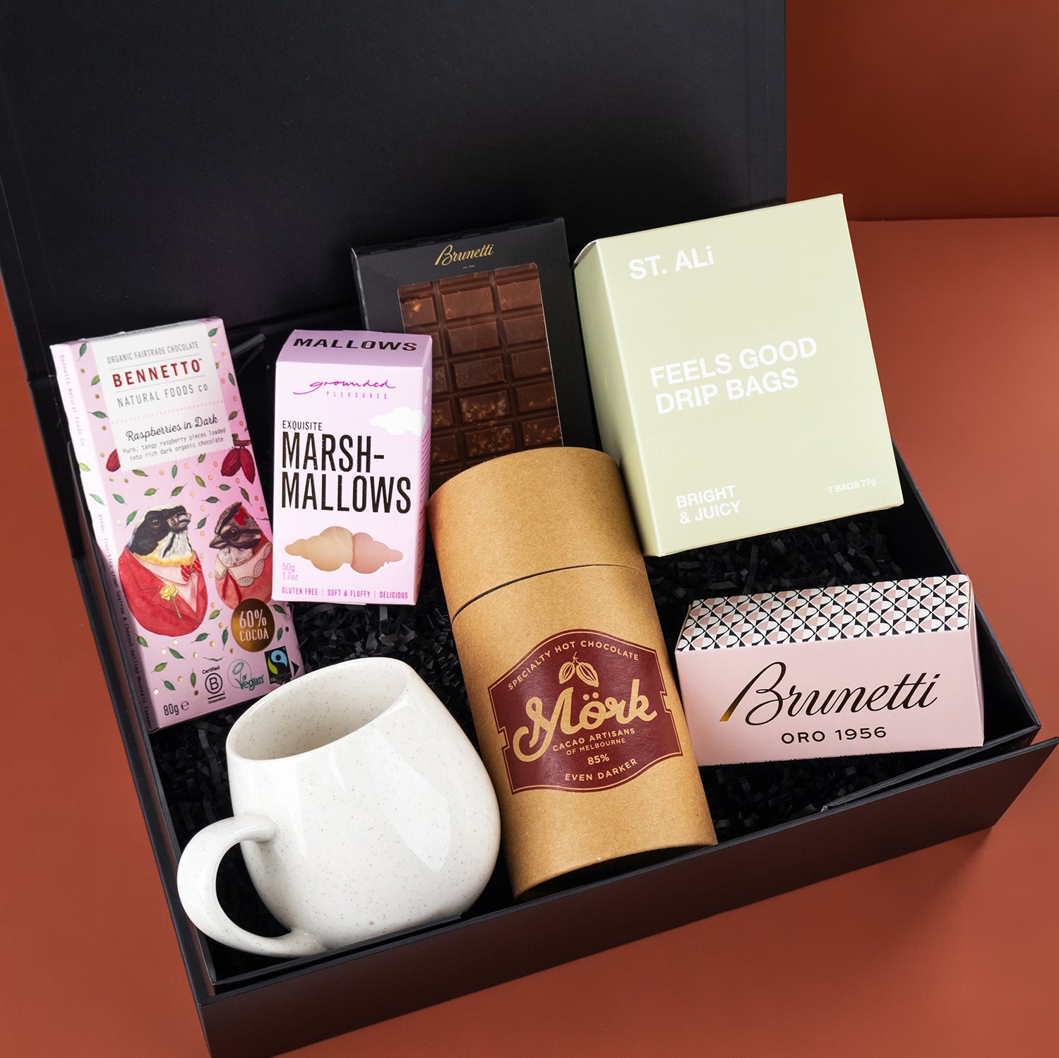 Black gift box with a white ceramic mug, marshmallows, chocolate bar, coffee canister, and a green box of tea bags.