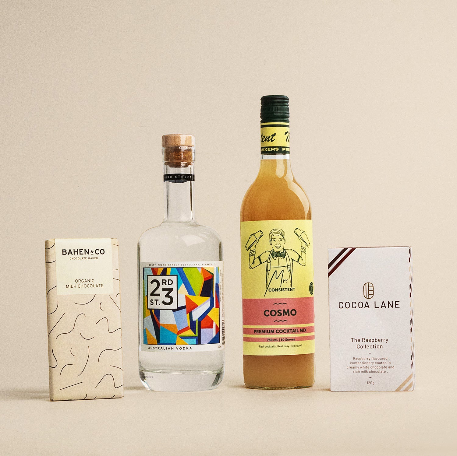 Cosmo Hamper with bottle of clear spirit, yellow Cosmo mixer, and two boxed gourmet snacks on beige background.