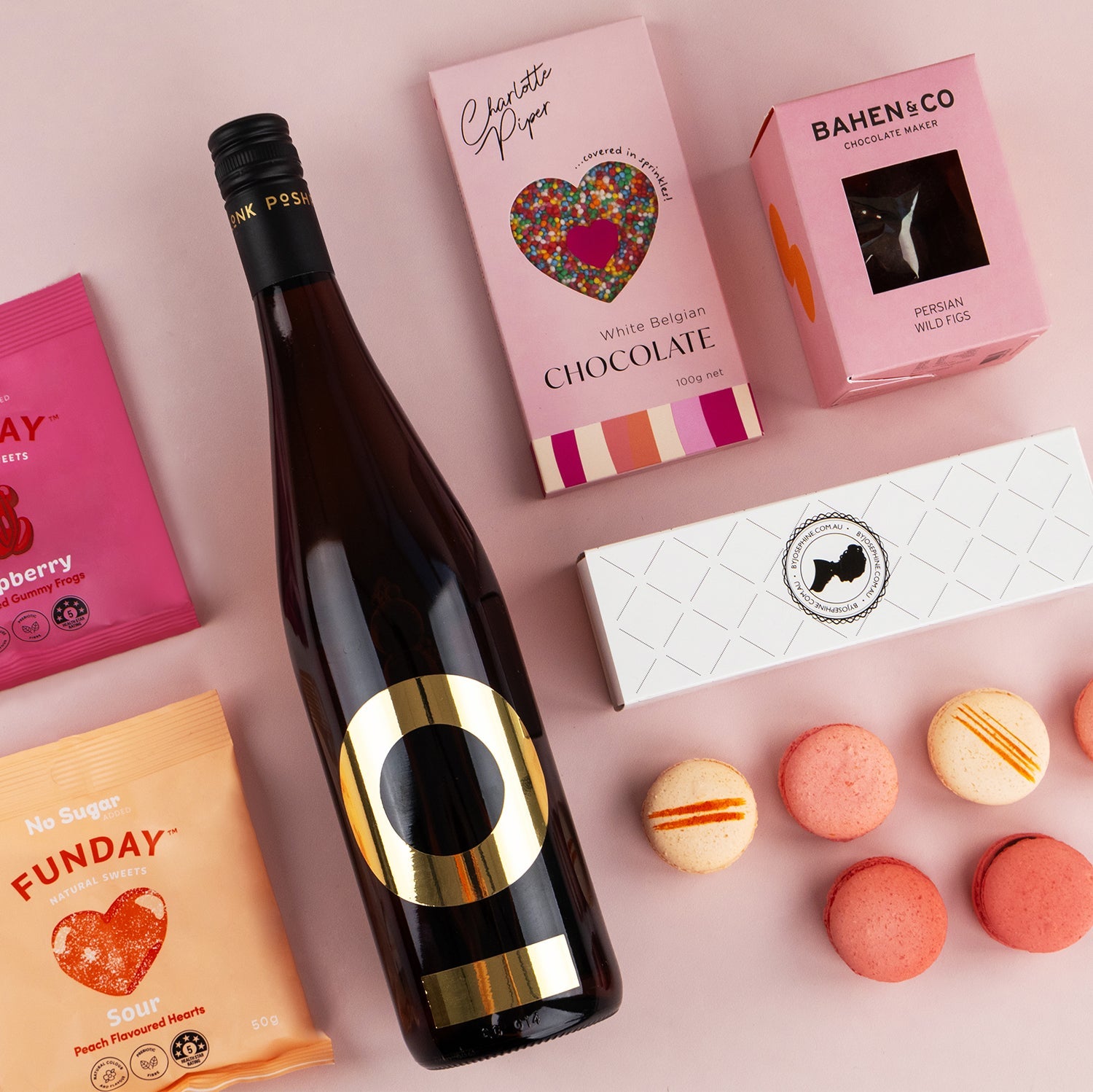 Bottle of Posh Plonk Pinot Grigio with pink boxes of chocolates and six assorted pink and orange macarons on white background.