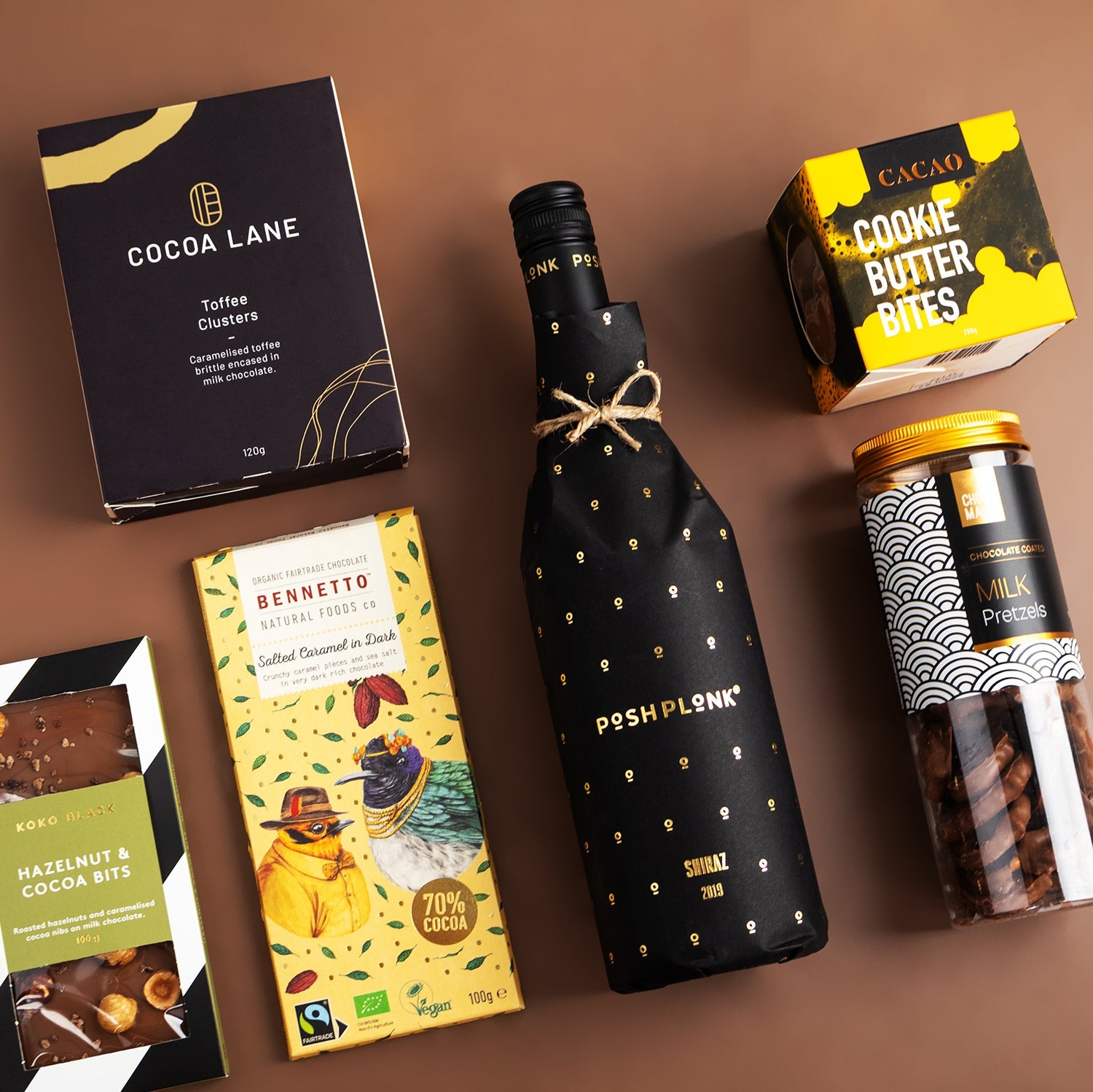 Posh Plonk Shiraz bottle wrapped with black paper and gold dots, surrounded by gourmet chocolate and cookie treats on a.
