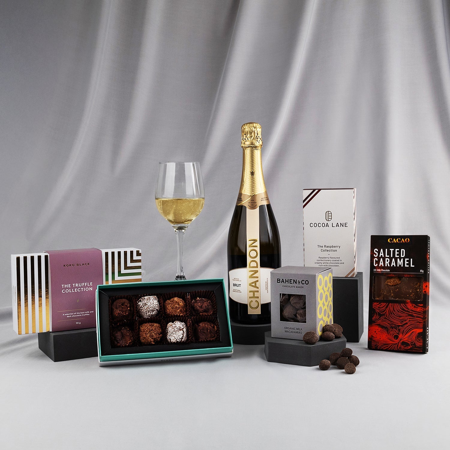 Chandon sparkling wine bottle with wine glass, assorted chocolates, salted caramel and white chocolate bars on gray satin.