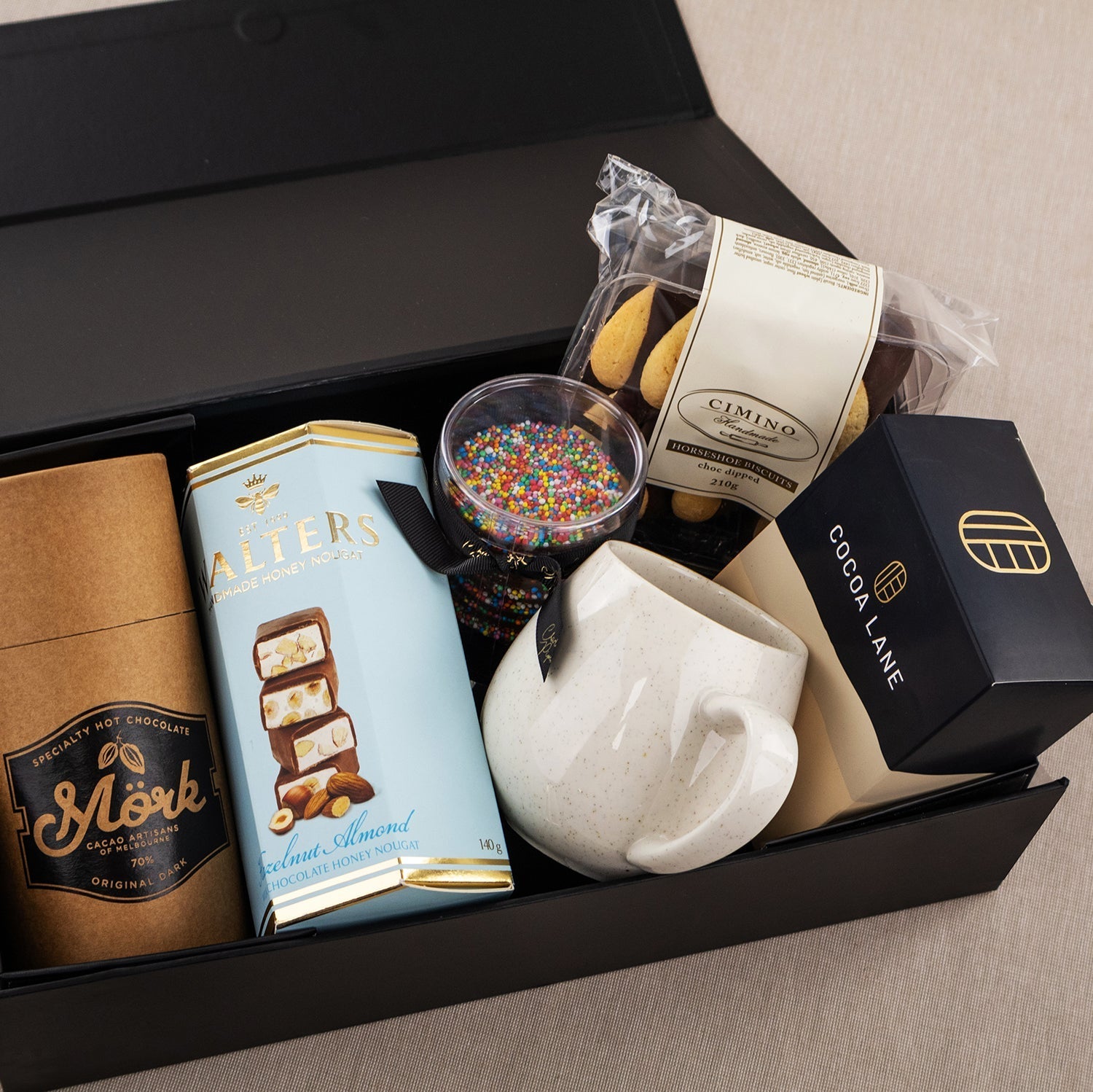 Black gift box with artisanal chocolates, coffee beans, a white ceramic mug, cookies, and colorful sprinkles in a clear cup.