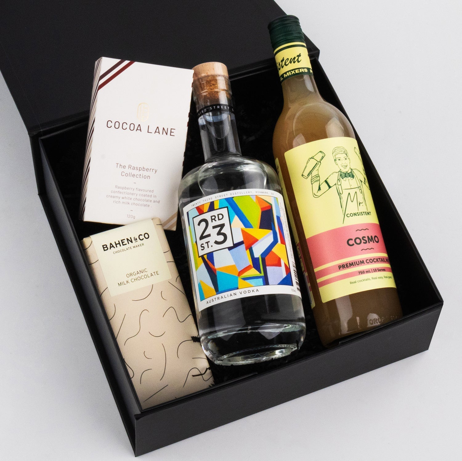 Black gift box with colorful liquor bottle, creamy liqueur bottle, and two beige and white chocolate bars.