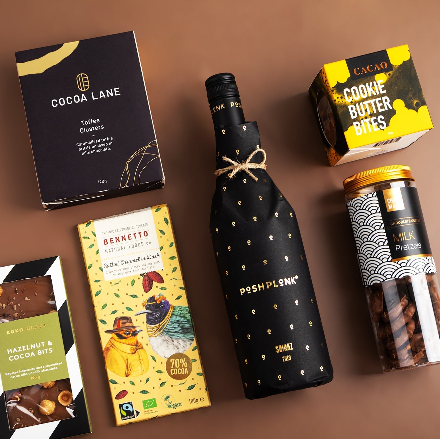 Posh Plonk Shiraz bottle wrapped in black with gold dots, chocolate bars, cookie butter bites, and jar of mixed nuts on.