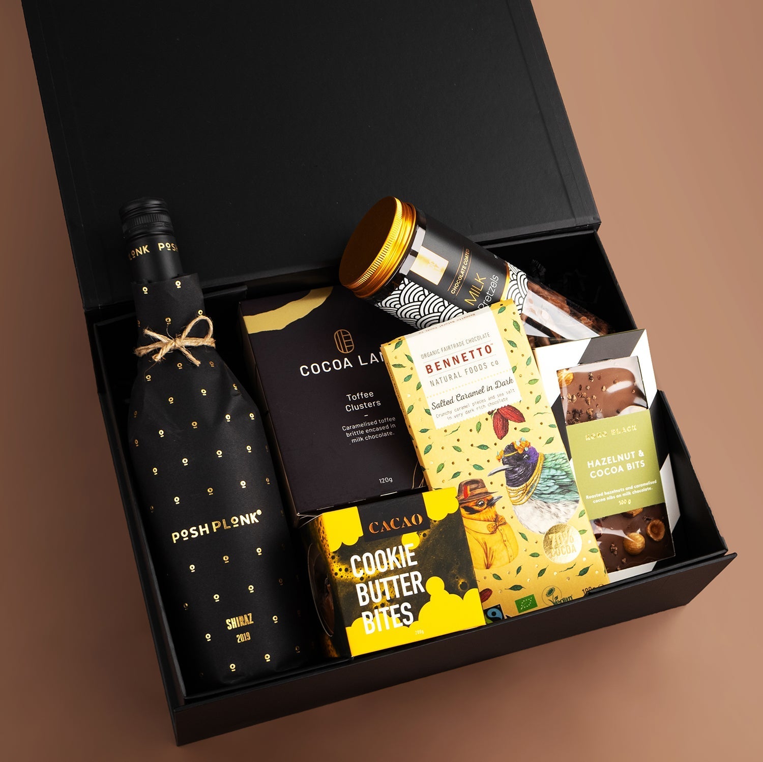Black bottle of Posh Plonk Shiraz with gold dots, dark chocolate, cookies, and gourmet treats in black gift box.