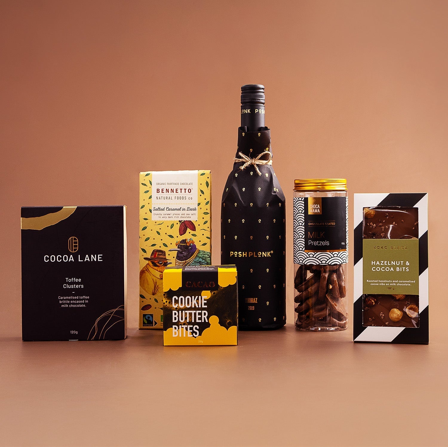 Elegant hamper featuring Posh Plonk Shiraz bottle, cookie butter bites, chocolate bars, and almond treats against a warm.