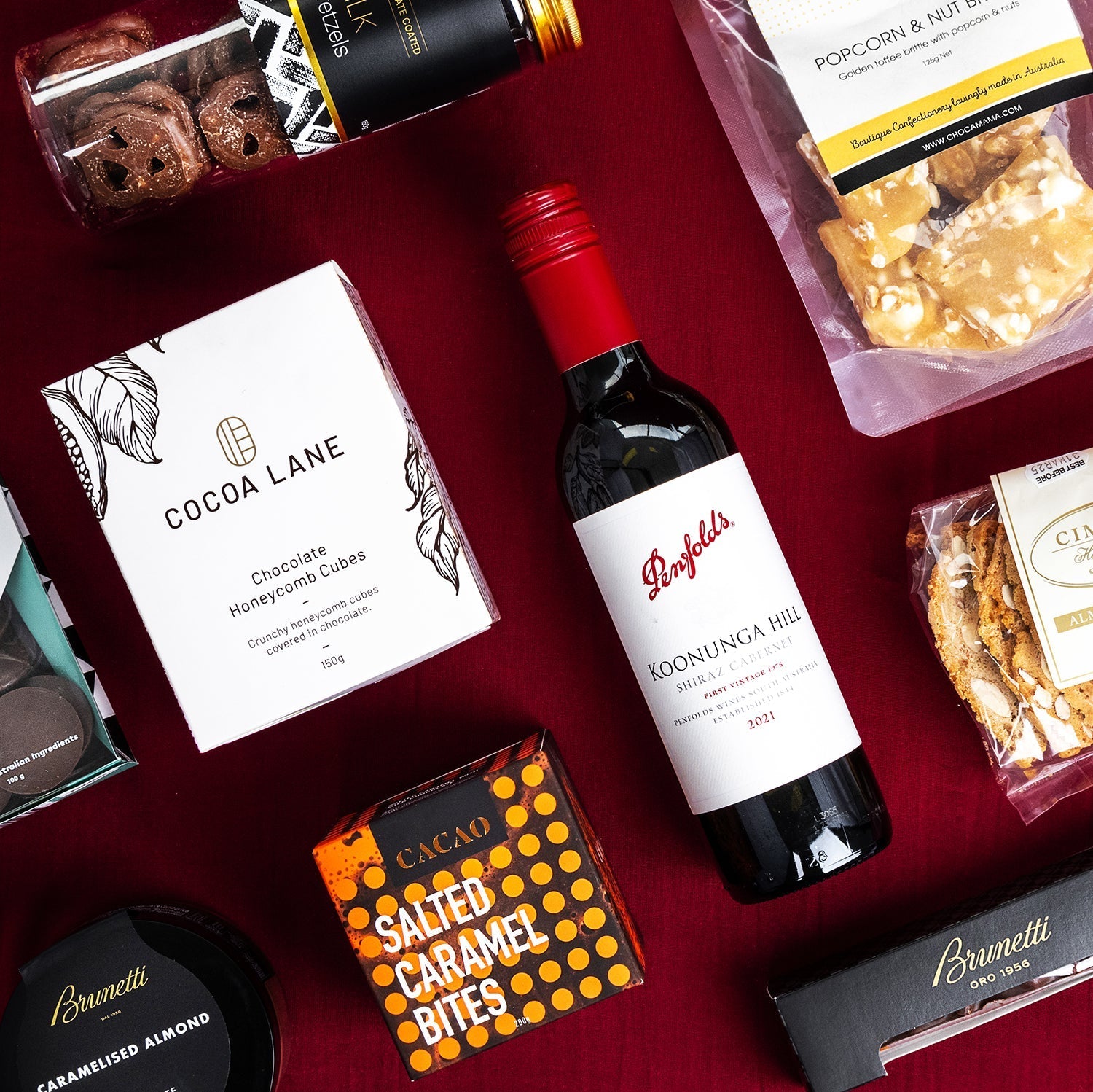 Red wine bottle surrounded by salted caramel bites, popcorn, chocolate, and gourmet treats on deep red background.