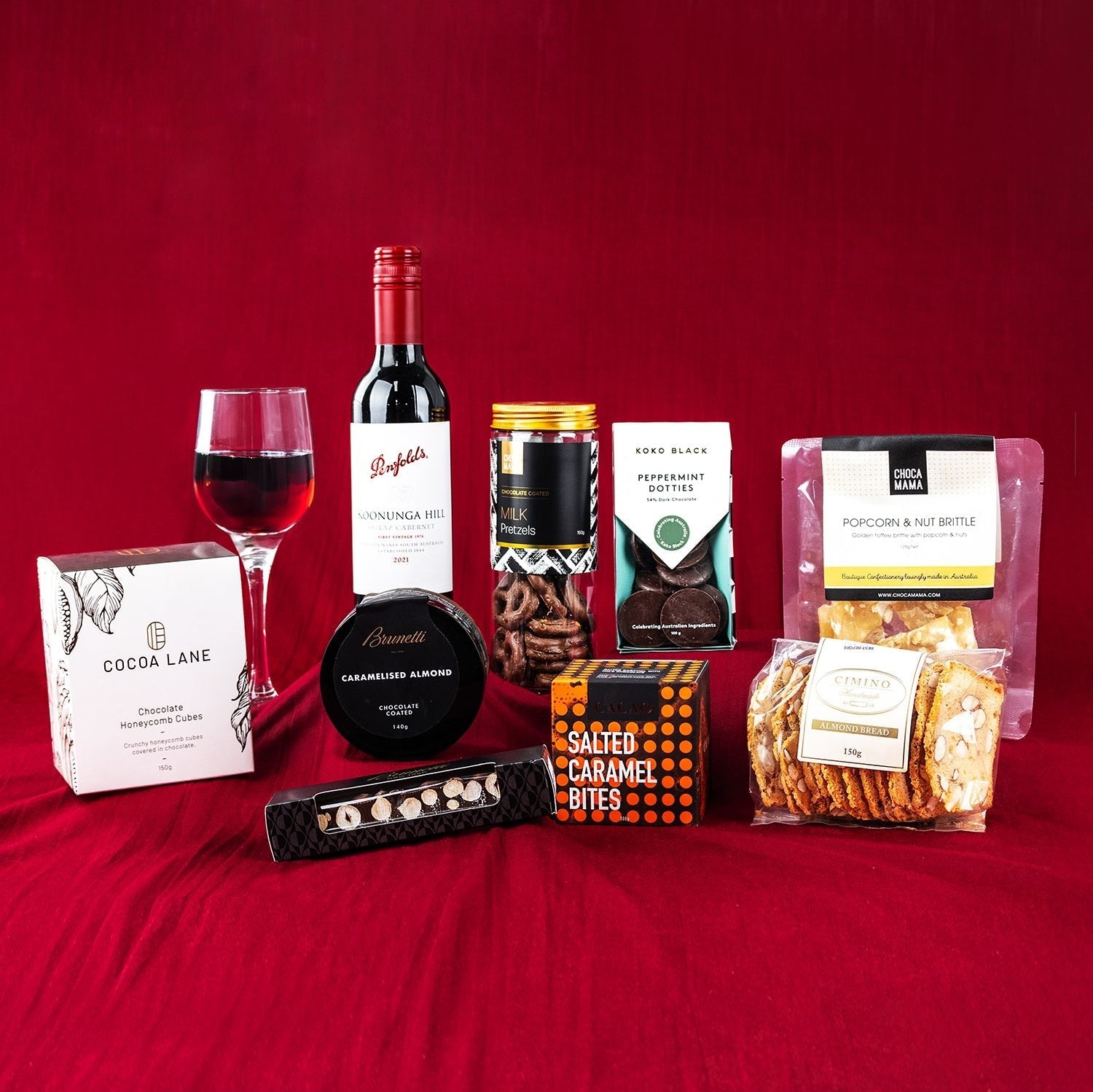Red Wine Indulgence Hamper featuring a bottle of red wine, filled wine glass, assorted chocolates, cookies, and gourmet.