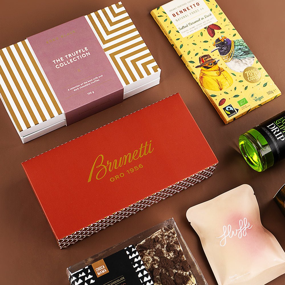 Luxury gift hamper with Brunetti biscotti and chocolate, artisan tea, and a small green bottle on brown background.
