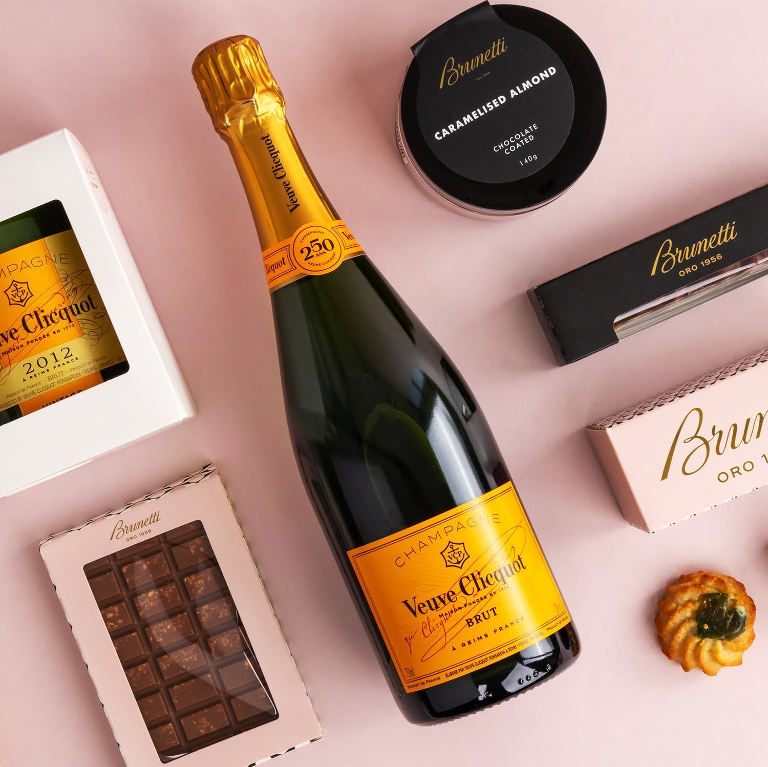 Bottle of Veuve Clicquot Yellow Label champagne with Brunetti branded chocolates and confectionery on a pink background.