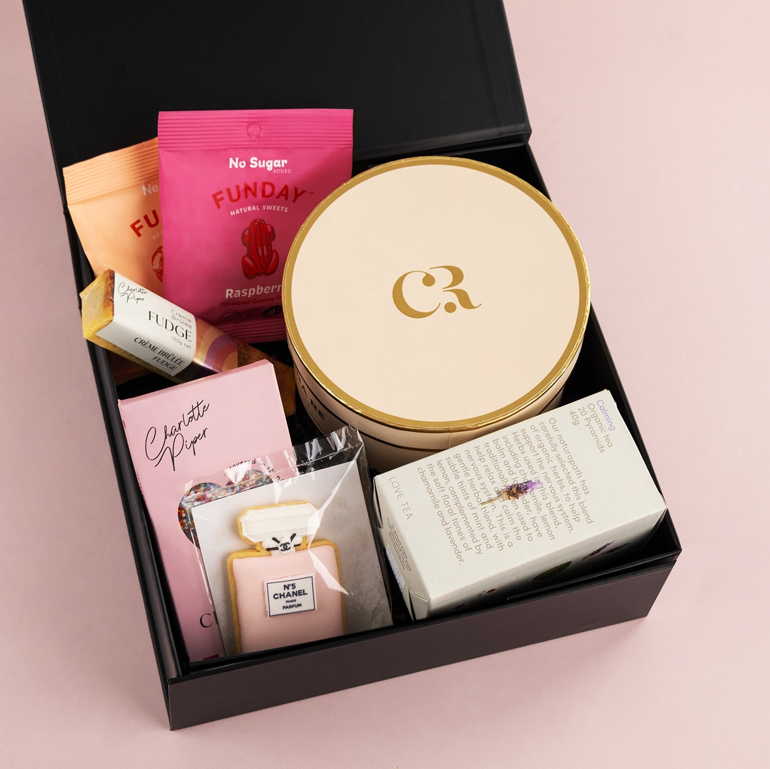 Black gift box with pink, gold, and cream packaged treats including tea and confectionery.
