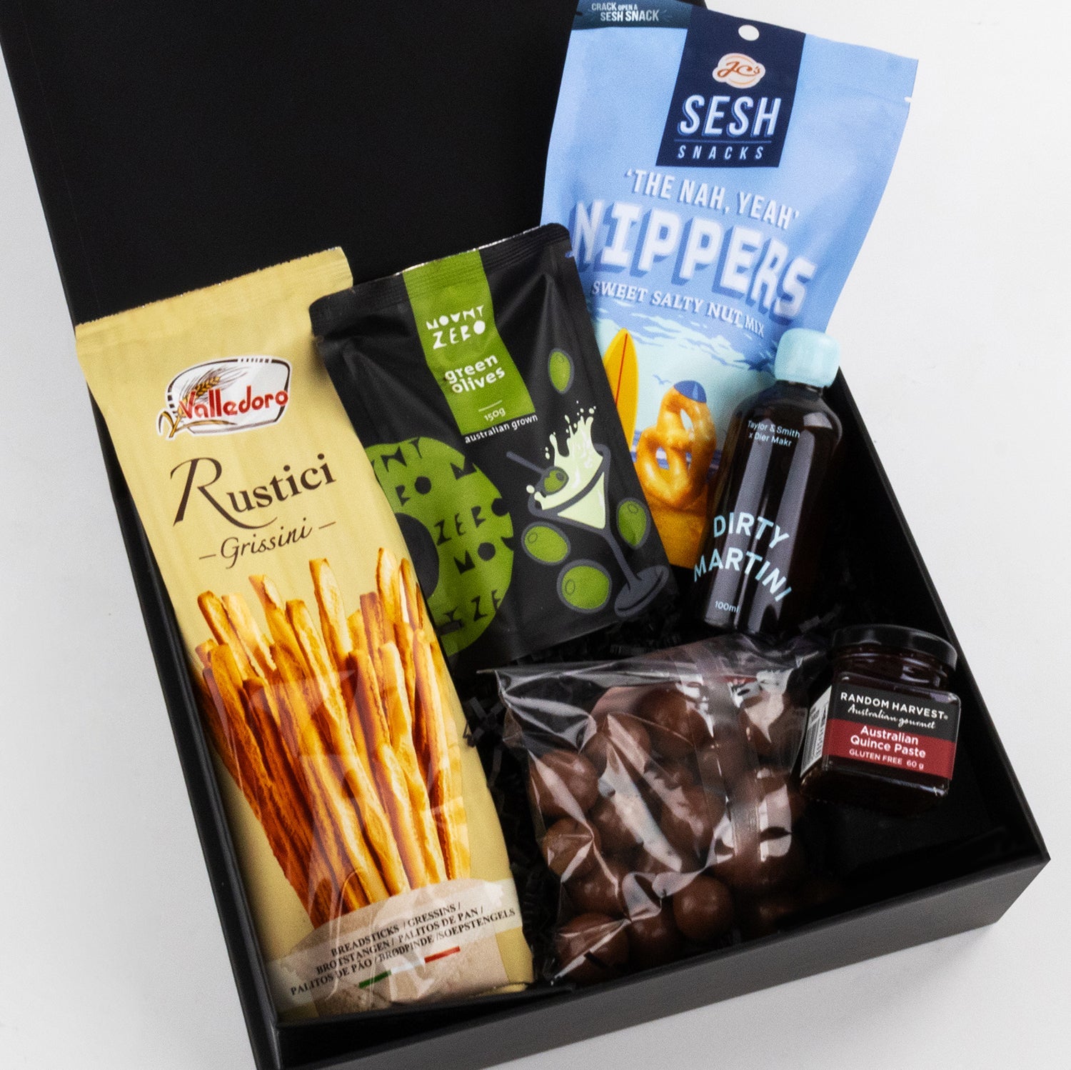 Black gift box with breadsticks, blue crisp bag, cocktail mixer bottle, chocolate pieces, and a jar of jam.