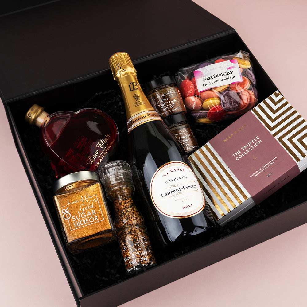 Black gift box containing heart-shaped bottle of red liquid, jar of golden sugar, pepper grinder, bottle of sparkling wine,.