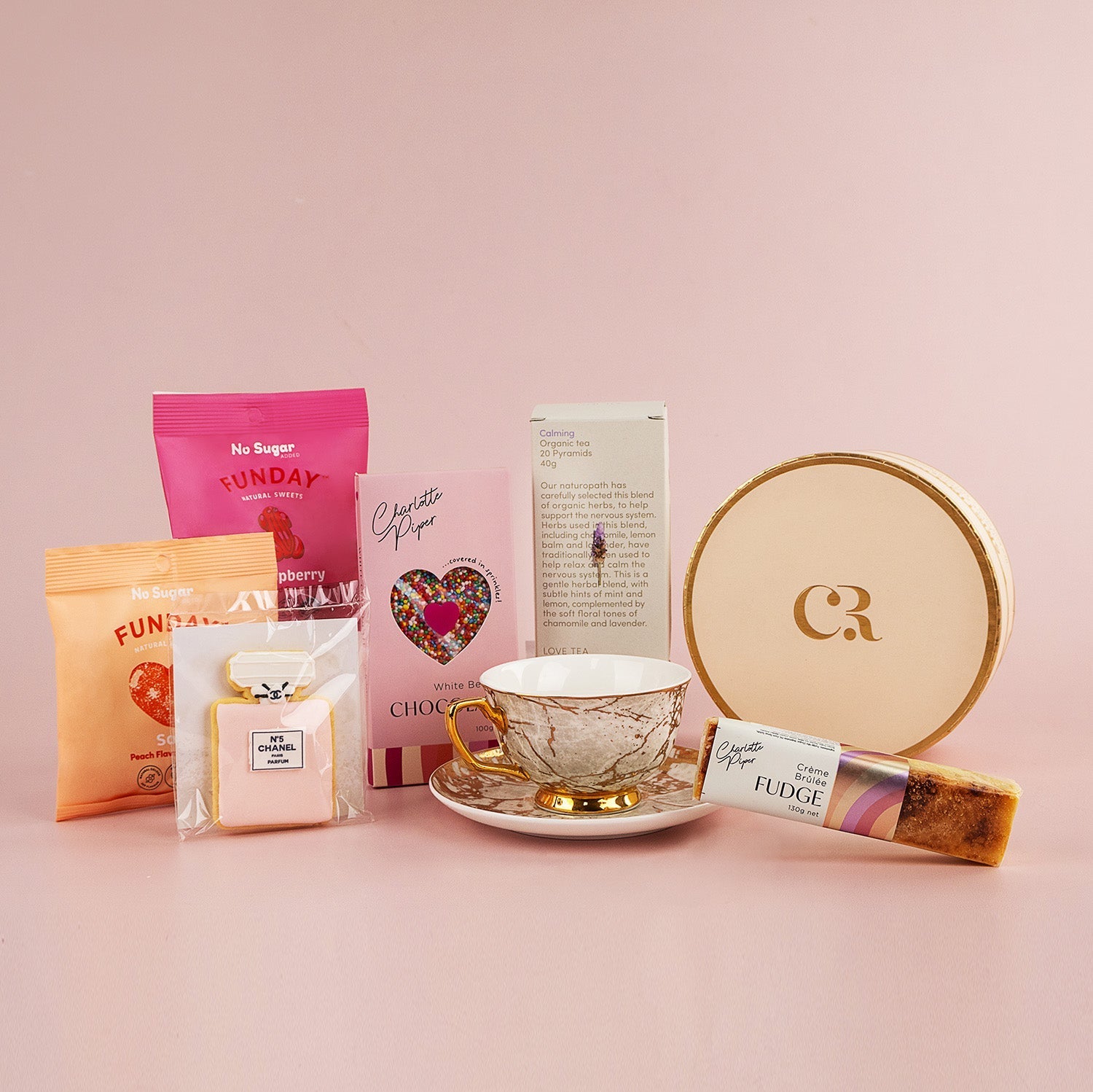 Celestite Hamper with white and gold teacup, round cream box, assorted pink and orange gourmet treats on blush background.