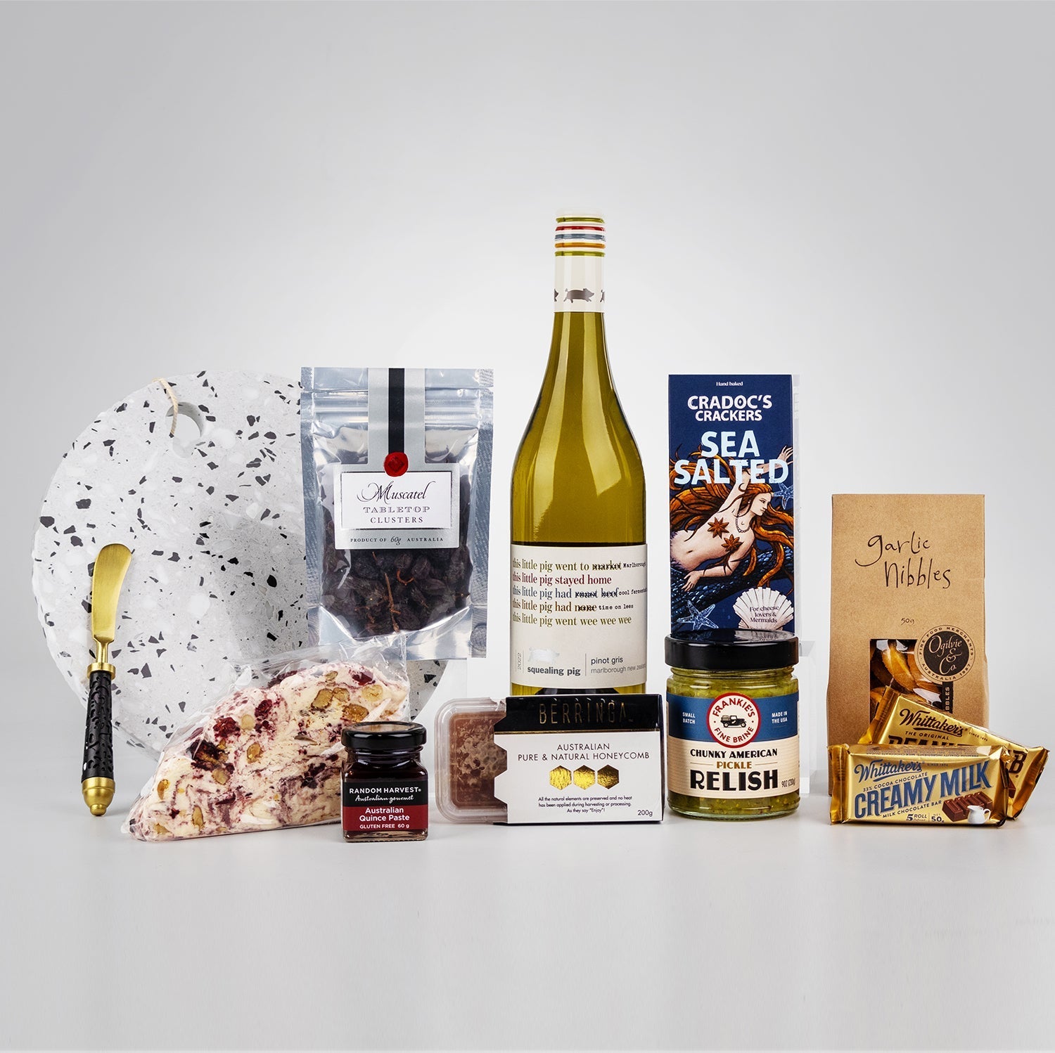 Charcuterie hamper with white wine, sea salt crackers, chocolate, cheese, relish, berry jam, and cheese knife on a white.