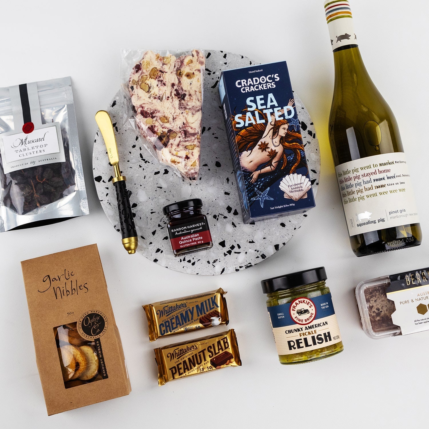 Charcuterie hamper with white wine, sea salted crackers, peanut slab, relish, chocolate pieces, and cheese on marble board.