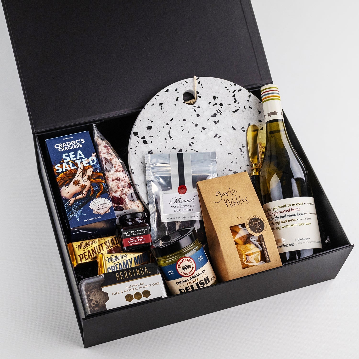 Black gift box with a bottle of white wine, terrazzo-patterned round cheese board, crackers, preserves, nuts, and chocolates.