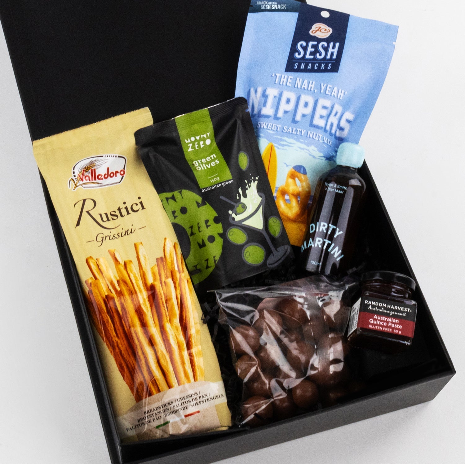 Black gift box with breadsticks, seaweed crackers, choco-covered nuts, Dirty Martini cocktail bottle, and balsamic glaze jar.