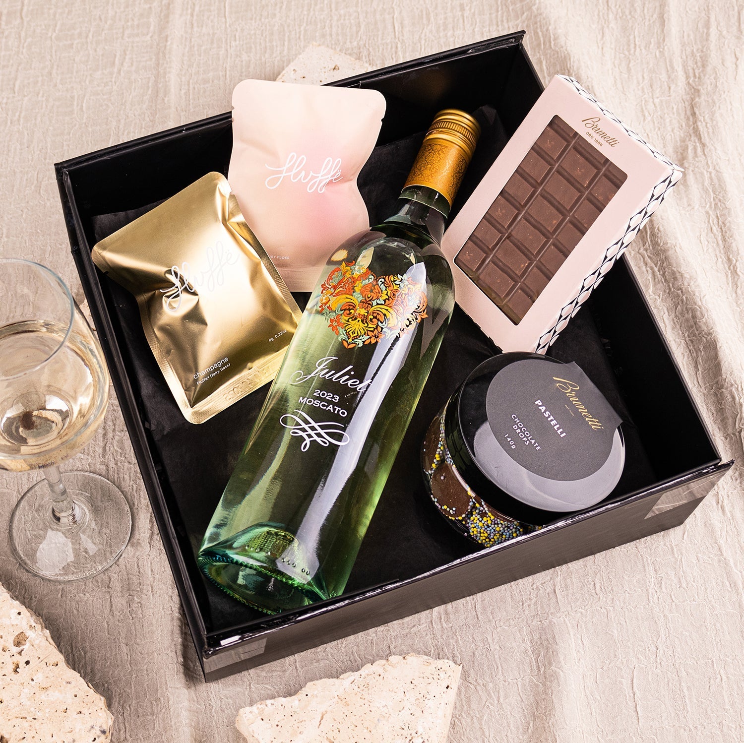 Black hamper box with a bottle of Juliet Moscato, gold-wrapped snack, chocolate bar, jar of preserves, and pink candle.